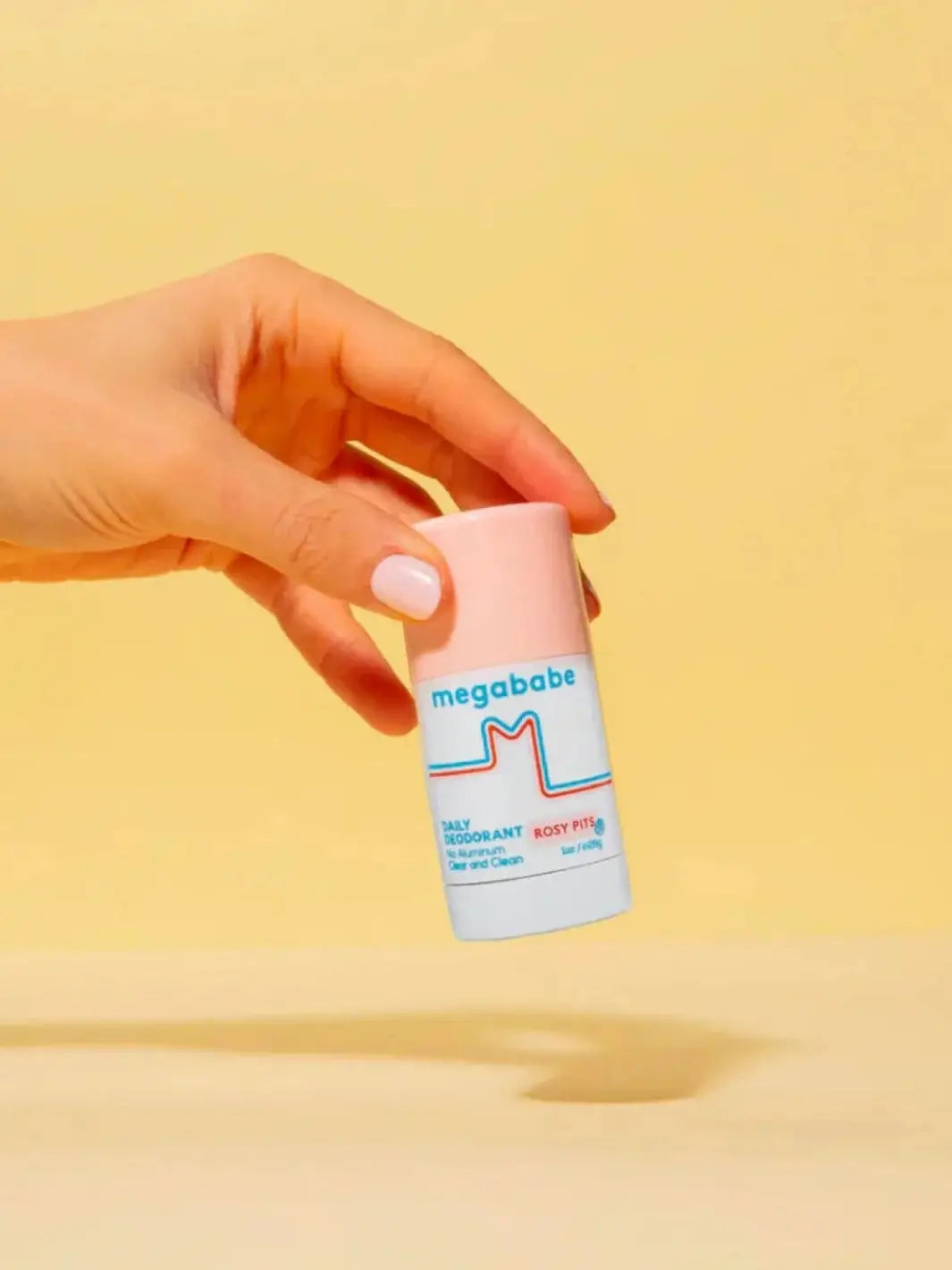A hand with a light pink manicure holds a travel-sized stick of Megababe Rosy Pits daily deodorant against a solid pastel yellow background | Twentyseven Toronto