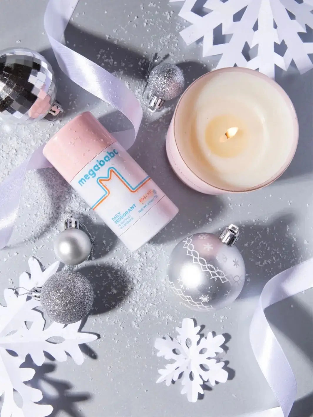 A top-down view captures a lit pink jar candle arranged amidst silver holiday ornaments, white paper snowflakes, and scattered faux snow to showcase the festive setting for the Mason Pearson Rosy Pits Scented Candle | Twentyseven Toronto