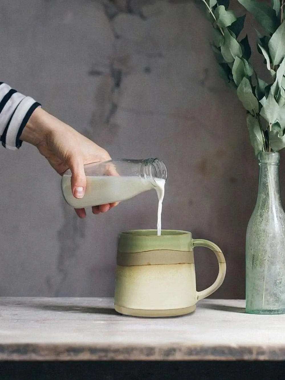A hand pours milk from a small glass bottle into an Abbott Collection Rustic Style Mug in Green resting on a textured tabletop | Twentyseven Toronto
