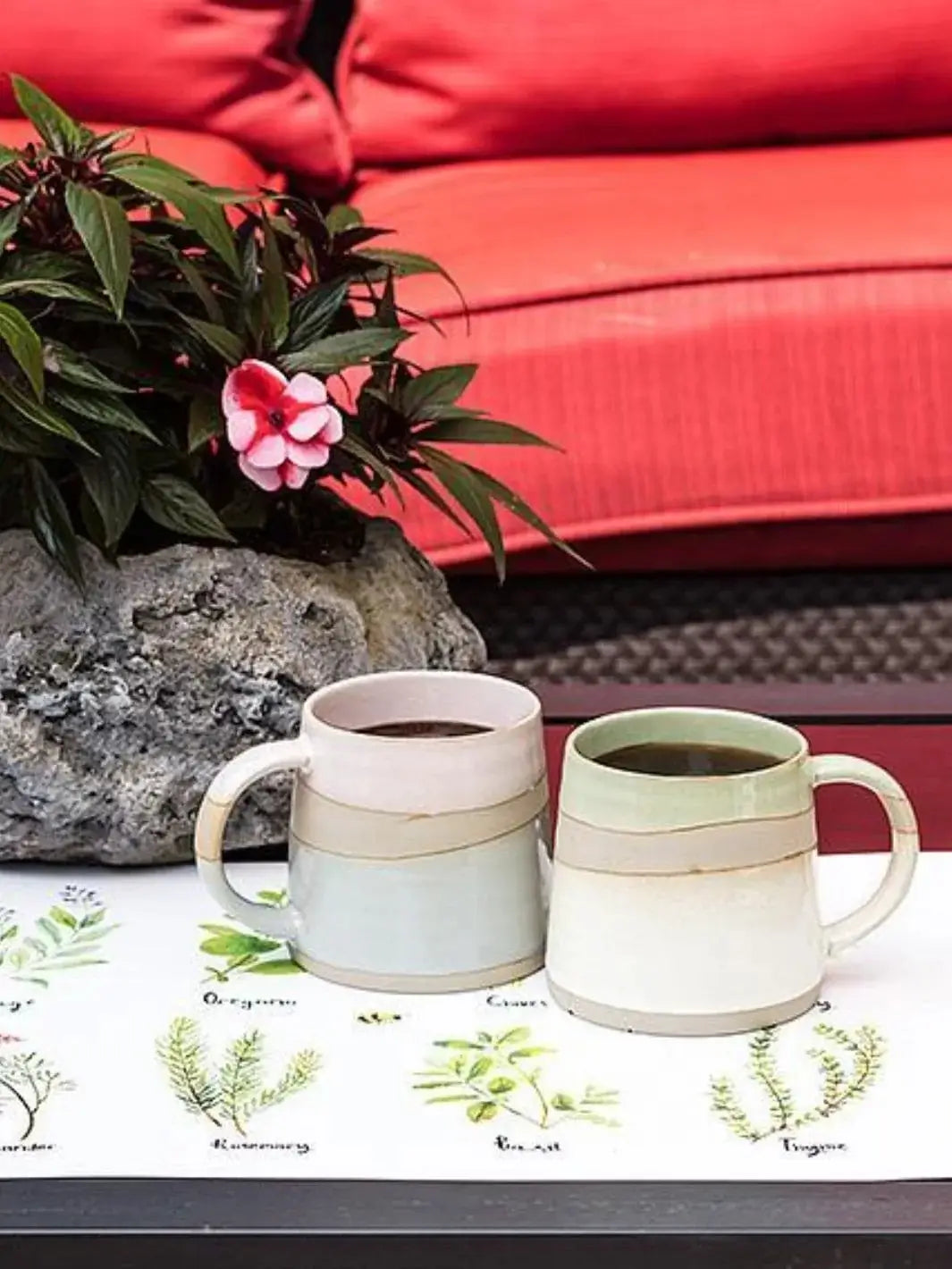 Two ceramic coffee mugs filled with dark coffee, including an Abbott Collection Rustic Style Mug in Green, sit on a botanical print mat next to a potted flowering plant | Twentyseven Toronto