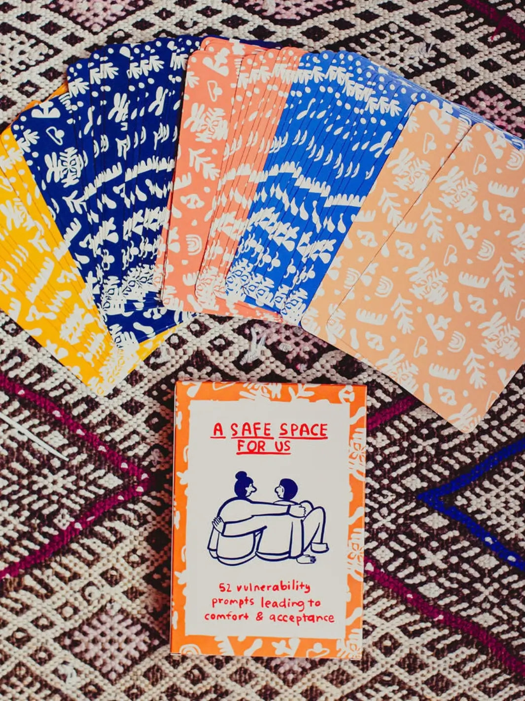 A fan of colorful, patterned cards spreads out above the box for the A Safe Space For Us Deck from People I've Loved resting on a textured, woven rug | Twentyseven Toronto