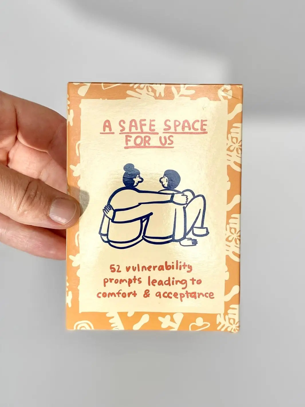 A hand holds the front packaging of the A Safe Space For Us Deck from People I've Loved, which features a minimalist illustration of two people hugging | Twentyseven Toronto