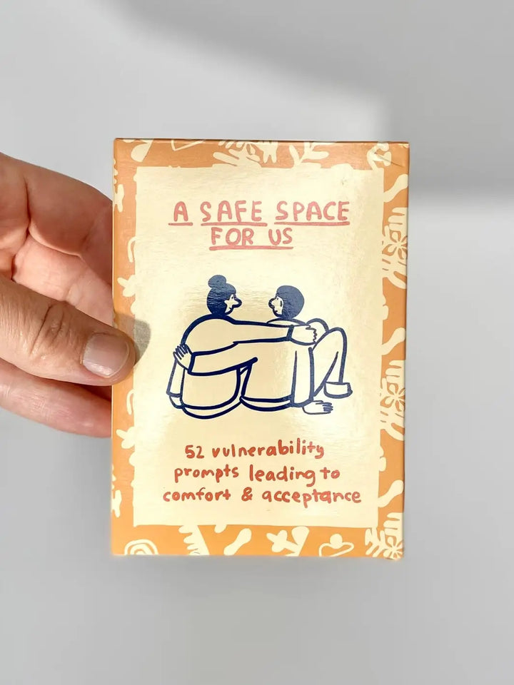 A hand holds the front packaging of the A Safe Space For Us Deck from People I've Loved, which features a minimalist illustration of two people hugging | Twentyseven Toronto
