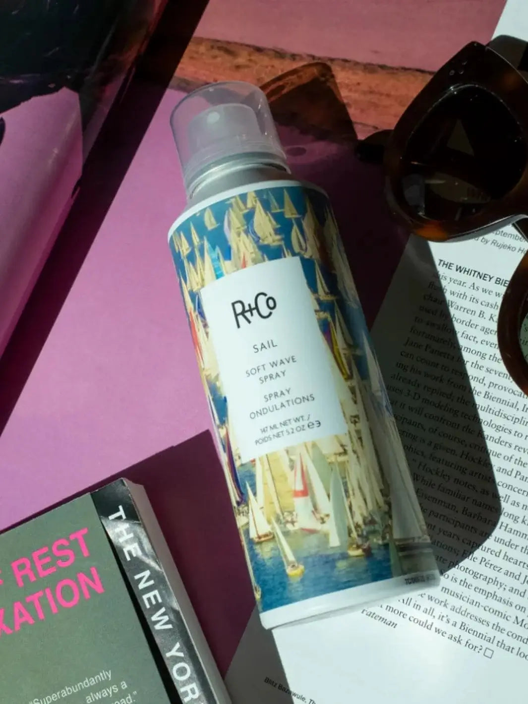 A bottle of R+Co SAIL Soft Wave Spray rests alongside sunglasses and printed reading materials on a vibrant pink background | Twentyseven Toronto