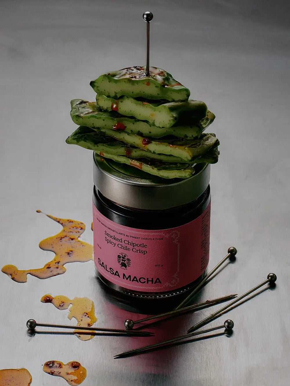 Sliced grilled nopales cactus stacked atop a jar of Flamingo Estate Salsa Macha Chile Crisp skewered with cocktail picks on a stainless steel surface | Twentyseven
