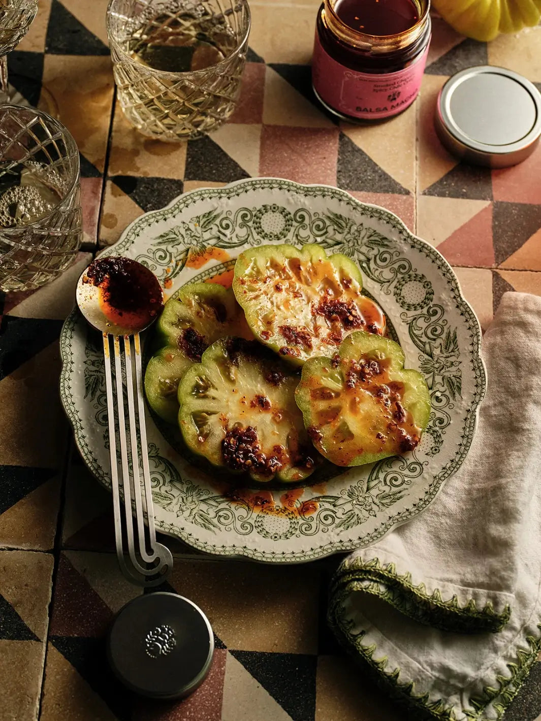 Green heirloom tomato slices drizzled with Flamingo Estate Salsa Macha Chile Crisp served on a vintage floral plate alongside cocktail glasses | Twentyseven