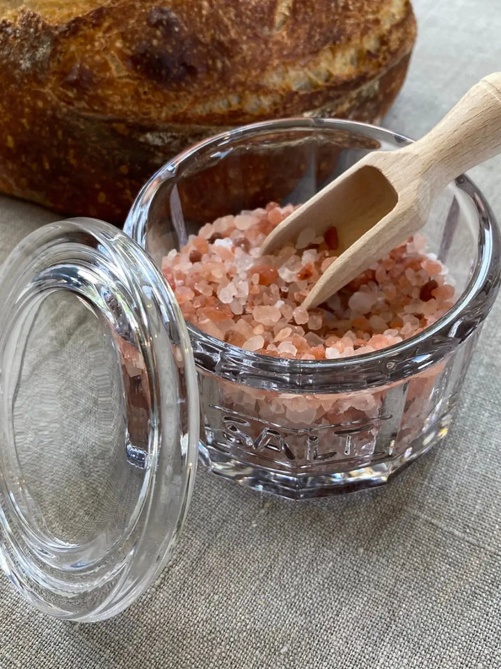 A clear glass Salt Pot w/ Scoop from Earth and Nest filled with pink Himalayan salt crystals and a small wooden scoop and an open lid next to a fresh loaf of bread | Twentyseven Toronto