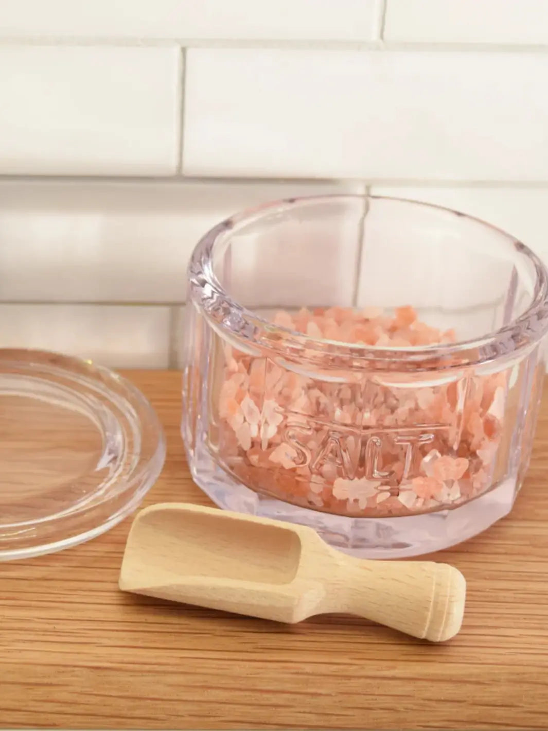 A clear glass Salt Pot w/ Scoop from Earth and Nest containing pink salt crystals on a wooden countertop next to its lid and a wooden scoop against a white background | Twentyseven Toronto