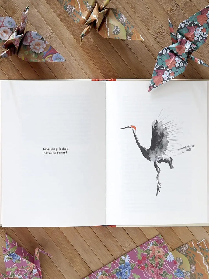 An open spread from Senbazuru: One Thousand Steps To Happiness, Fold By Fold by Michael James Wong displays a crane illustration and the quote "Love is a gift that needs no reward." The book is surrounded by colorful floral origami cranes on a wooden background | Twentyseven Toronto