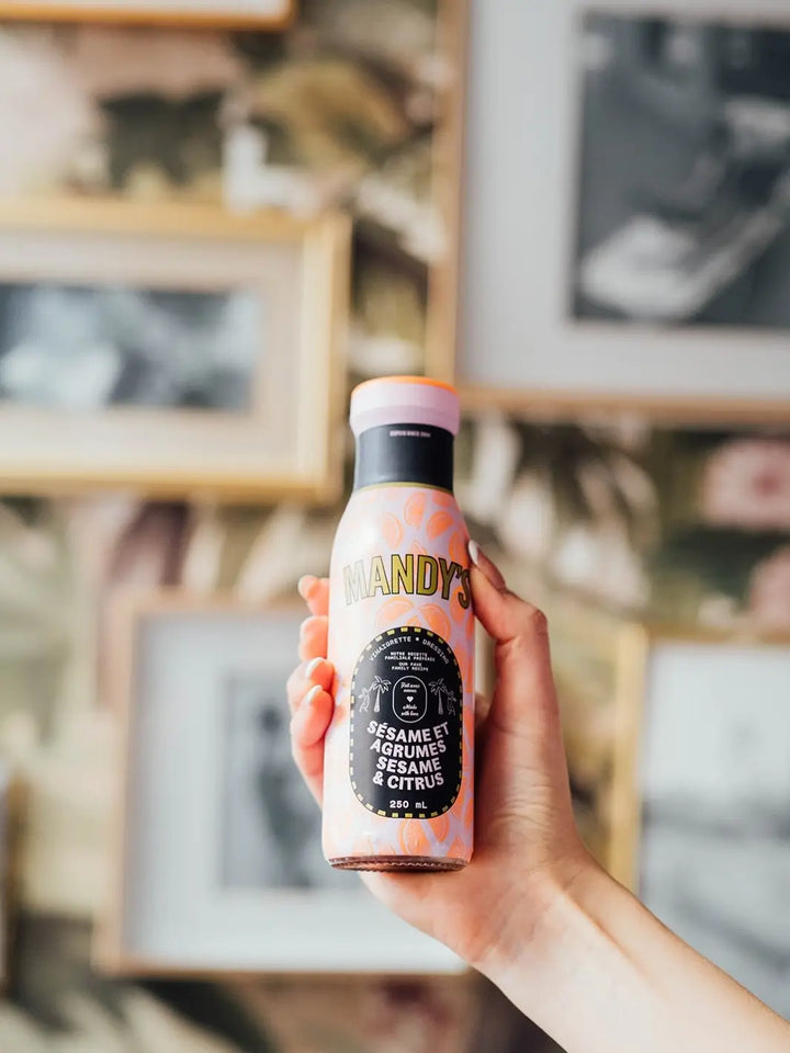 A person's hand holds up a 250 mL bottle of Mandy's Sesame Citrus dressing against a softly blurred background of framed wall art | Twentyseven Toronto