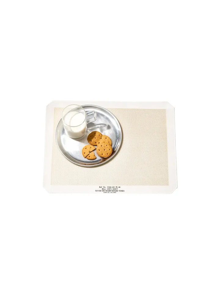 A top-down shot shows a metal plate filled with cookies and a clear mug of milk resting on a white Silicone Placemat from Puebco featuring industrial reference text printed along its bottom edge | Twentyseven Toronto