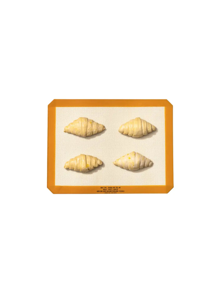 Four croissants are arranged on an orange-bordered Silicone Placemat from Puebco, demonstrating its dual-purpose functionality as both a dining accessory and a baking liner | Twentyseven Toronto