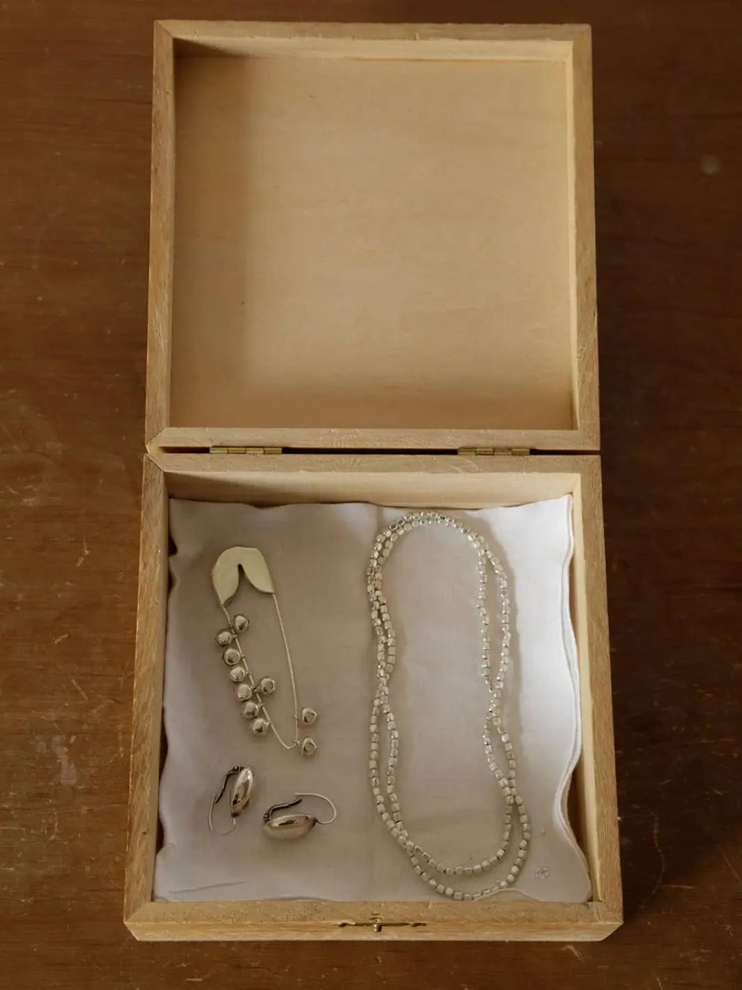 An open wooden jewelry box reveals a Fog Linen Work Silver Safety Pin Bell Small resting on a white cloth alongside a matching beaded necklace and small silver earrings | Twentyseven Toronto