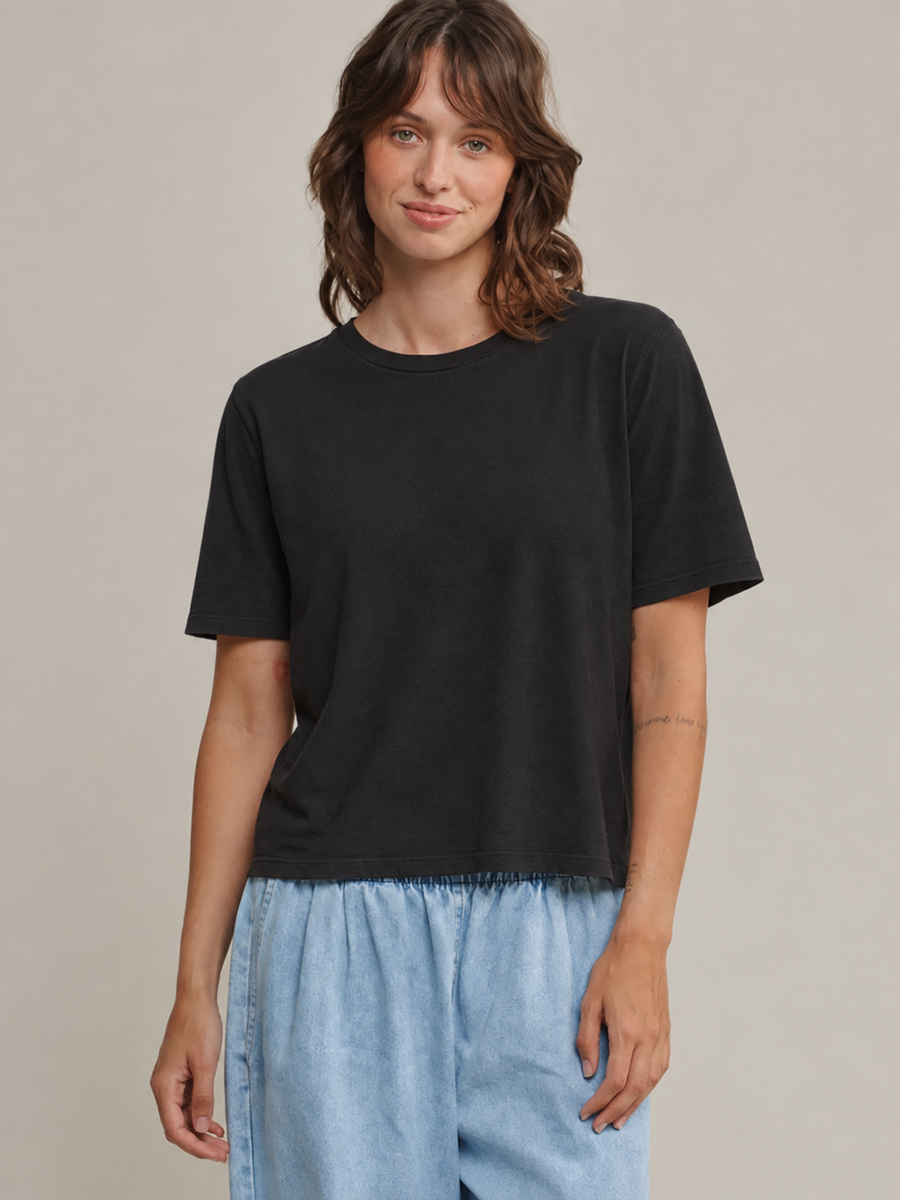 Front view of a brunette model posing in the black Jungmaven Silverlake Cropped Tee and light blue pants against a beige backdrop | Twentyseven 