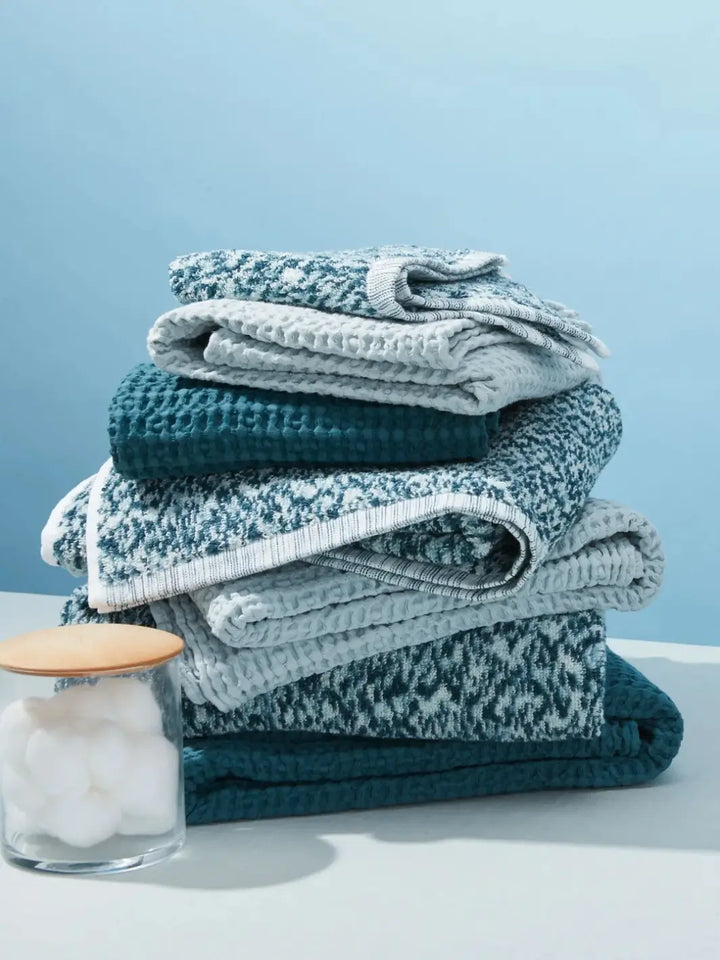 A neatly stacked pile of blue textiles features a light-colored bath sheet from the Simple Waffle Towel - Sky from Hawkins New York range layered with darker teal and patterned towels | Twentyseven Toronto