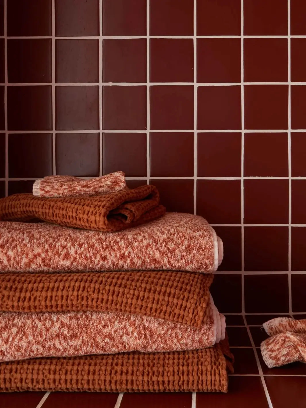 Stack of rich bath linens mixes patterned terry cloth with the textured layers of the Simple Waffle Towel - Terracotta from Hawkins New York against a backdrop of dark reddish-brown square tiles | Twentyseven Toronto