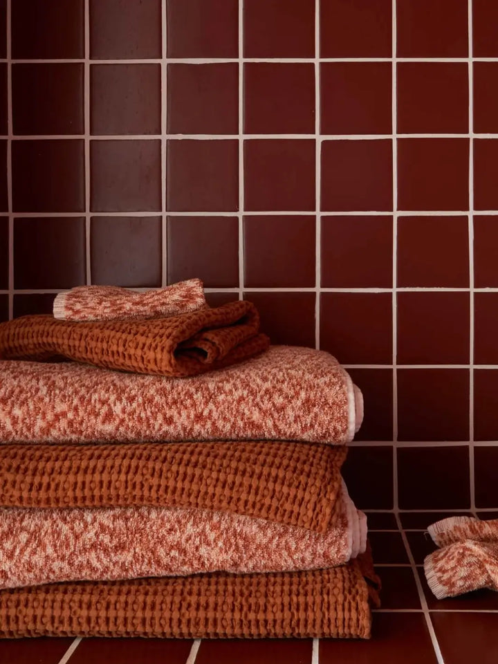 Stack of rich bath linens mixes patterned terry cloth with the textured layers of the Simple Waffle Towel - Terracotta from Hawkins New York against a backdrop of dark reddish-brown square tiles | Twentyseven Toronto