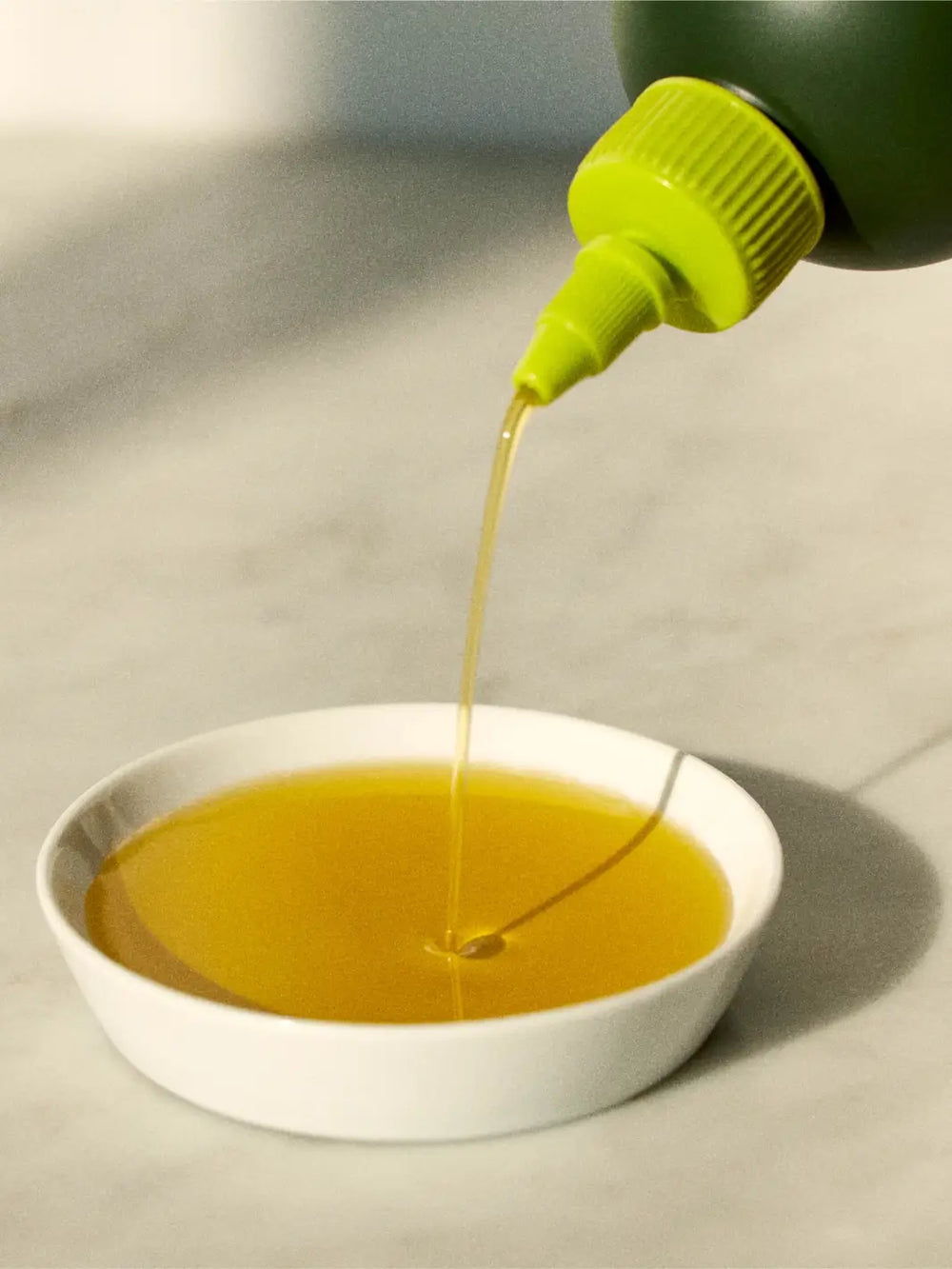 Green bottle of Graza 'Sizzle' pouring oil into a white bowl on a light surface | Twentyseven Toronto