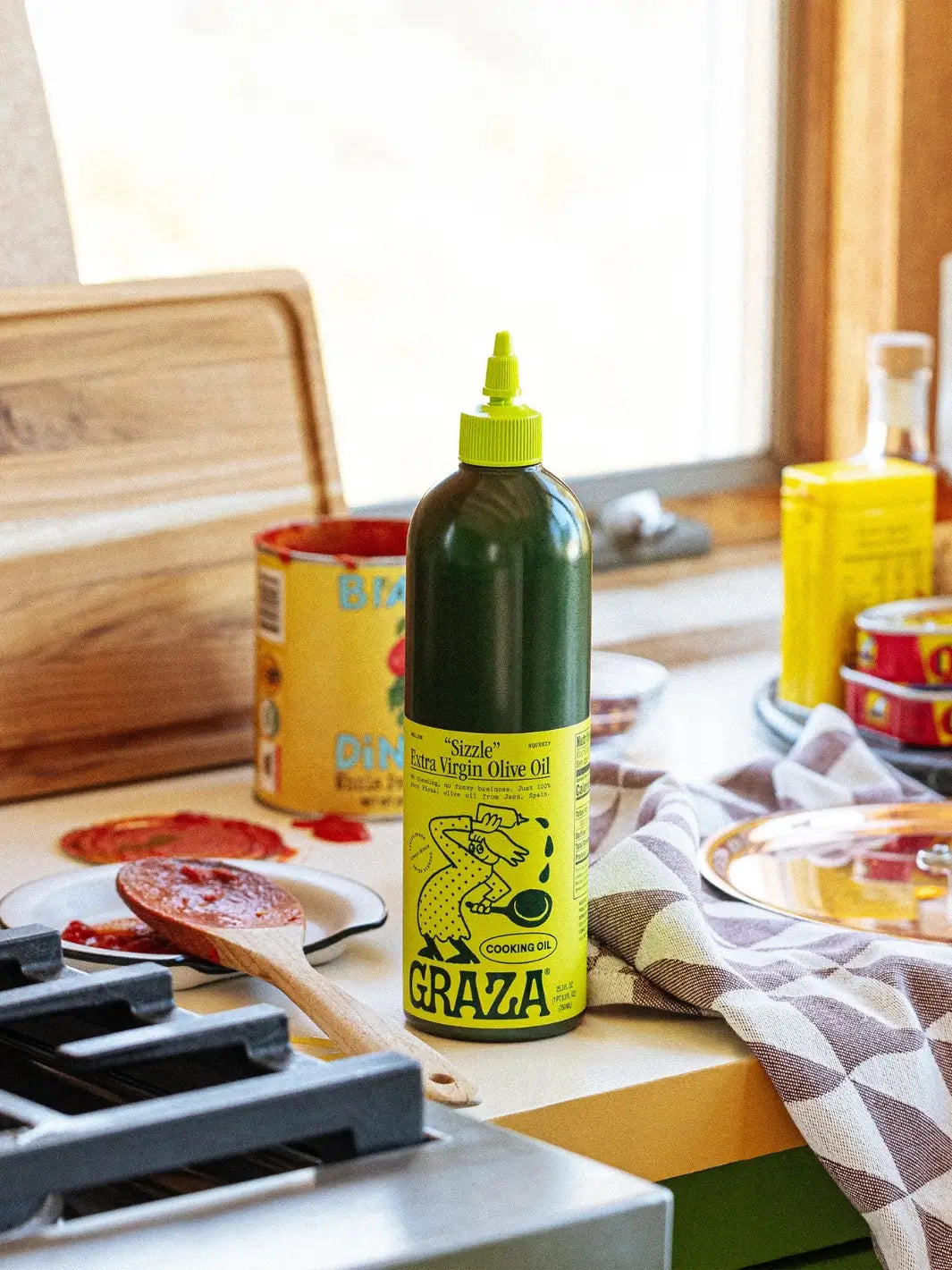 Bottle of Graza 'Sizzle' olive oil on a kitchen counter with food items in the background | Twentyseven Toronto