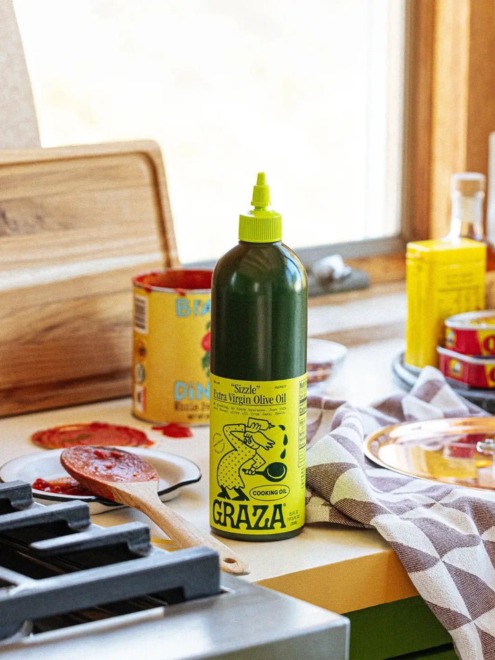 Bottle of Graza 'Sizzle' olive oil on a kitchen counter with food items in the background | Twentyseven Toronto