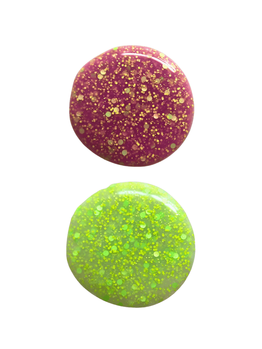 Two circular swatches of glittery 'Skunk Dreams' nail polish by Death Valley Nails, one pink and one green, on a transparent background | Twentyseven Toronto