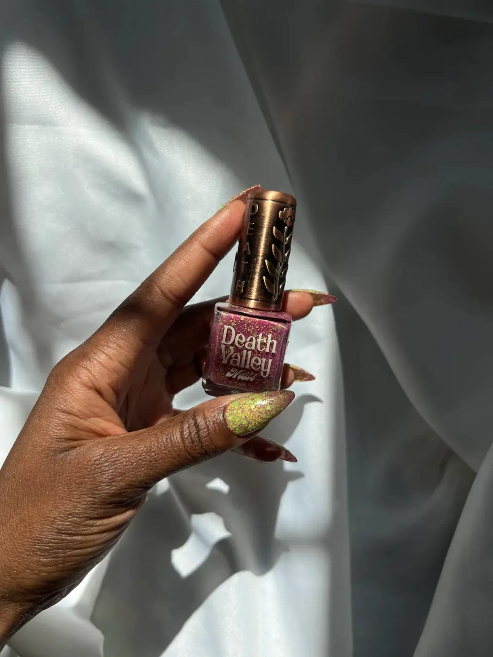 Hand holding a small bottle of 'Skunk Dreams Death Valley Nails' nail polish showing glittery green nails against a white background | Twentyseven Toronto