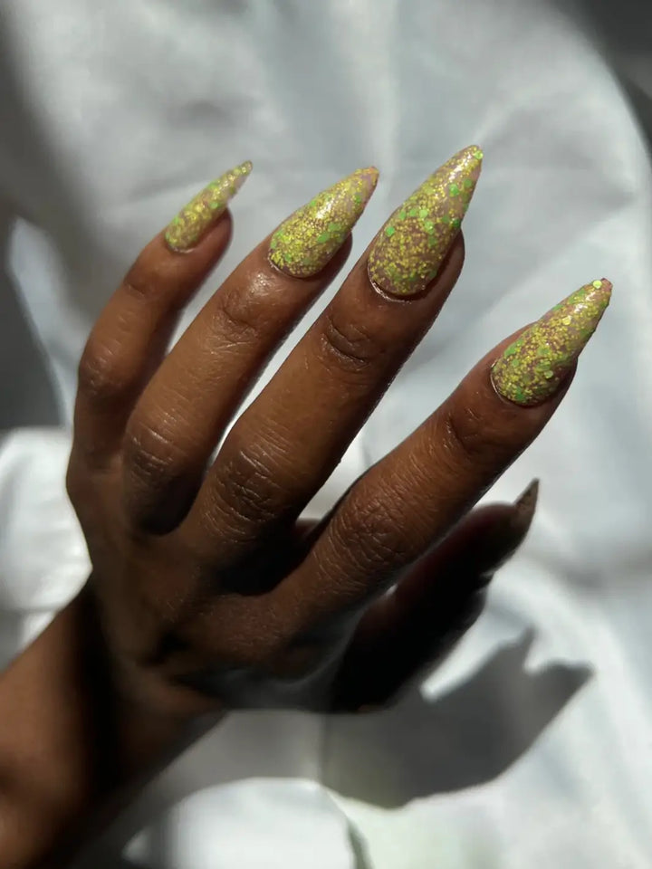 Hand with long glittery green nails, using 'Skunk Dreams' nail polish by Death Valley Nails on a white background | Twentyseven Toronto