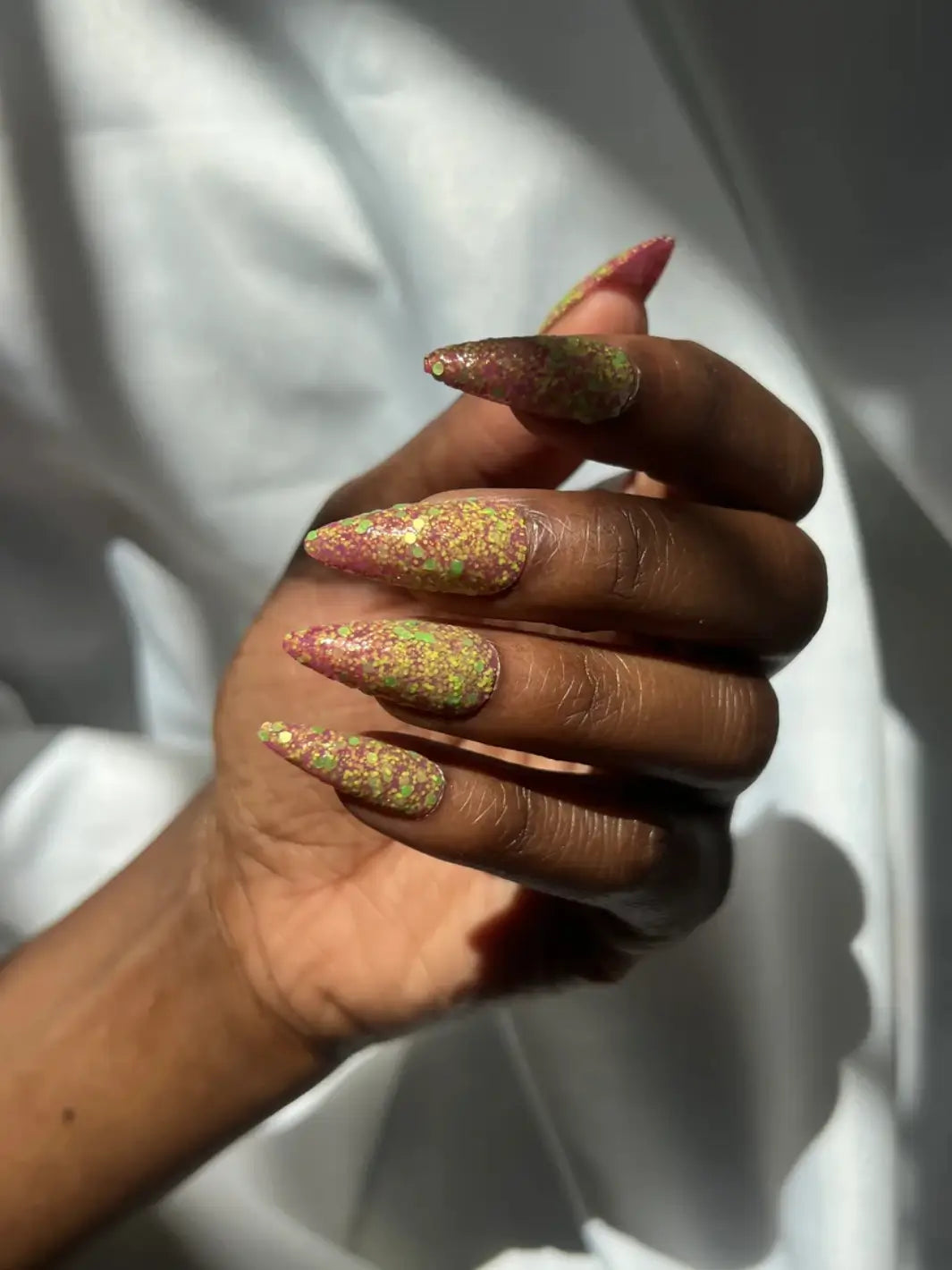 Hand with long glittery pink and green nails, using 'Skunk Dreams' nail polish by Death Valley Nails on a white background | Twentyseven Toronto