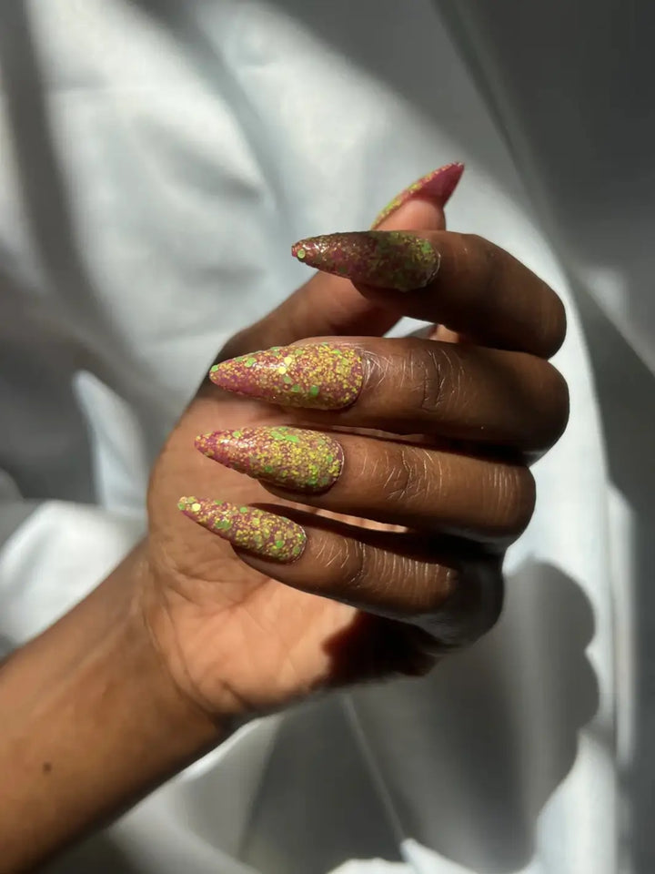 Hand with long glittery pink and green nails, using 'Skunk Dreams' nail polish by Death Valley Nails on a white background | Twentyseven Toronto