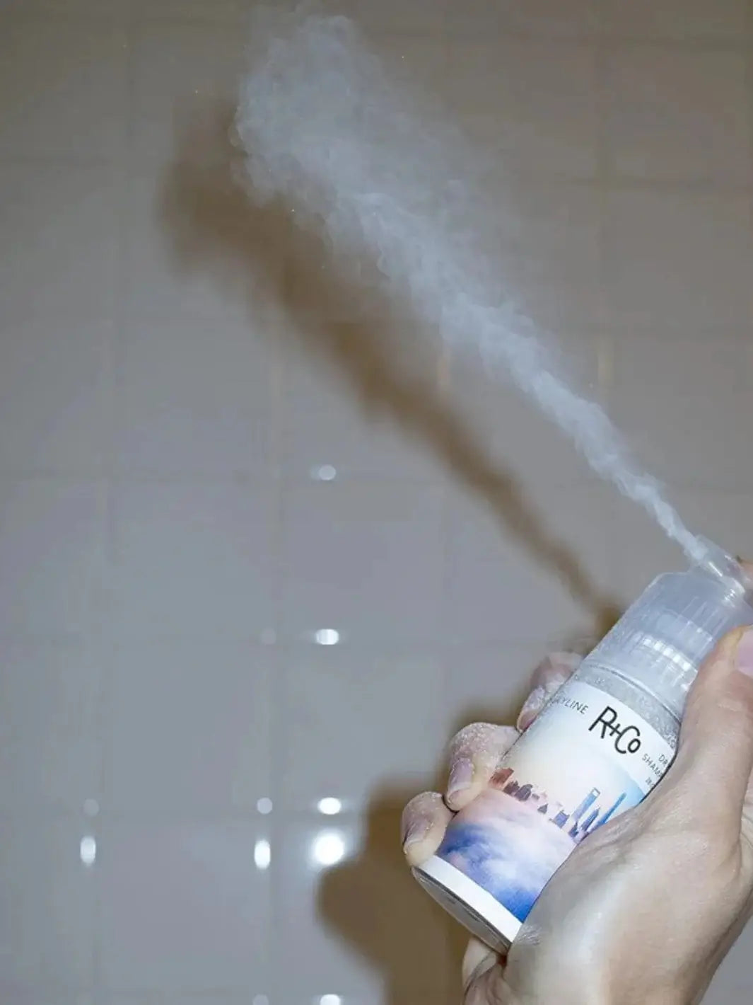 A hand pumps a bottle of R+Co SKYLINE Dry Shampoo Powder, releasing a dramatic cloud of fine white powder into the air | Twentyseven Toronto