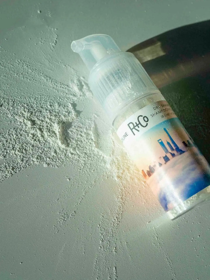 A bottle of R+Co SKYLINE Dry Shampoo Powder lies on a textured surface surrounded by scattered fine white powder | Twentyseven Toronto