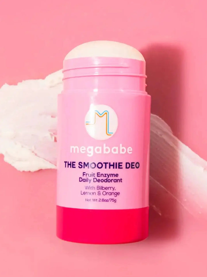 An opened pink deodorant stick rests vertically on a solid pink surface against a white swipe of product, highlighting the creamy texture of Megababe The Smoothie Deo | Twentyseven Toronto