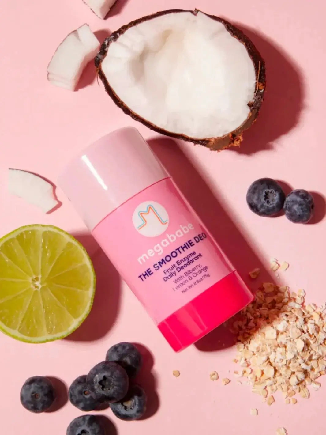 A pink deodorant stick lies flat on a light pink surface surrounded by a halved coconut, fresh lime, blueberries, and oats to emphasize the natural ingredients of Megababe The Smoothie Deo | Twentyseven Toronto