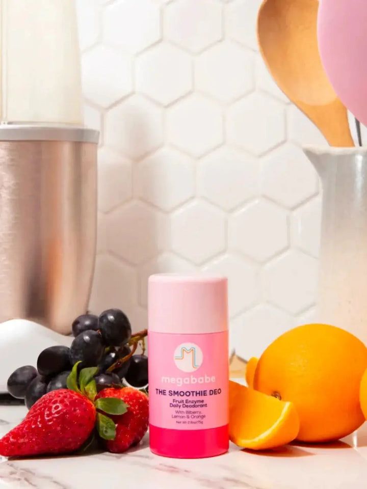 A pink deodorant stick is displayed on a kitchen counter alongside fresh strawberries, grapes, and oranges in front of a blender to showcase the fruit enzyme inspiration behind Megababe The Smoothie Deo | Twentyseven Toronto