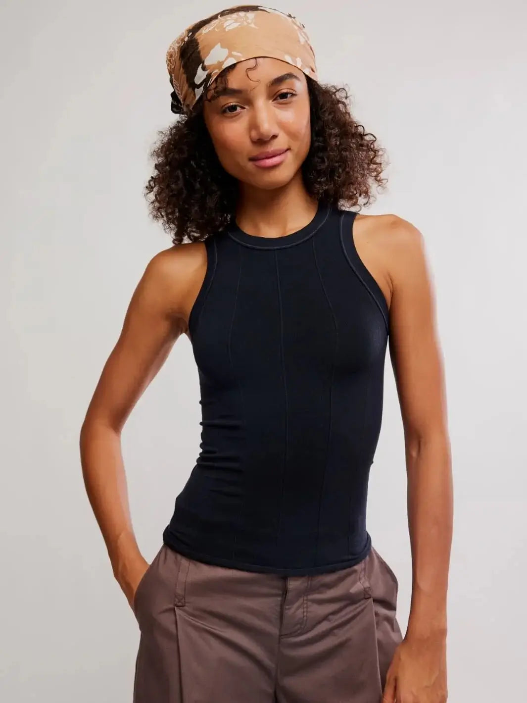A studio front view showcases a model wearing the ribbed, high-neck Free People So Soft Seamless Modal Tank in Black paired with brown trousers and a patterned hair scarf | Twentyseven Toronto