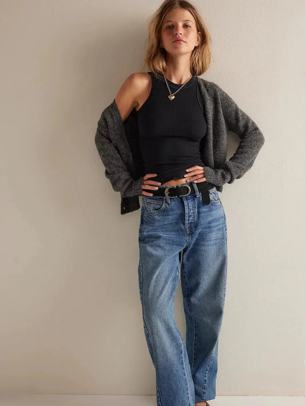 A full-length studio shot displays a model posing in the Free People So Soft Seamless Modal Tank in Black styled with blue jeans, a black belt, and an open charcoal grey cardigan | Twentyseven Toronto