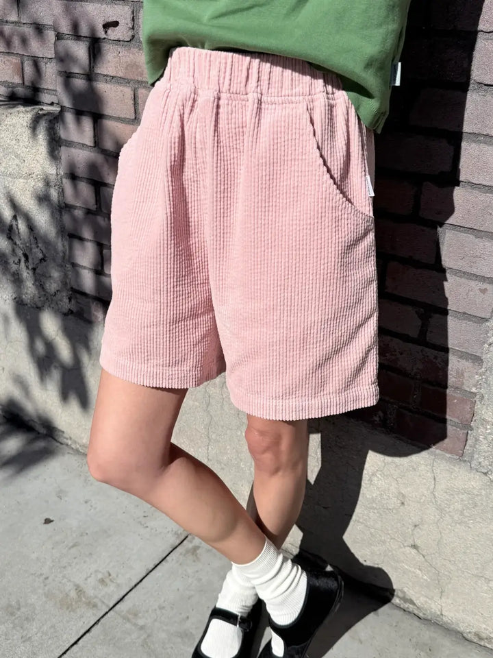 Person wearing pink 'Soft Corduroy City Shorts' in 'Baby Pink' and a green top from Le Bon Shoppe against a brick wall | Twentyseven Toronto