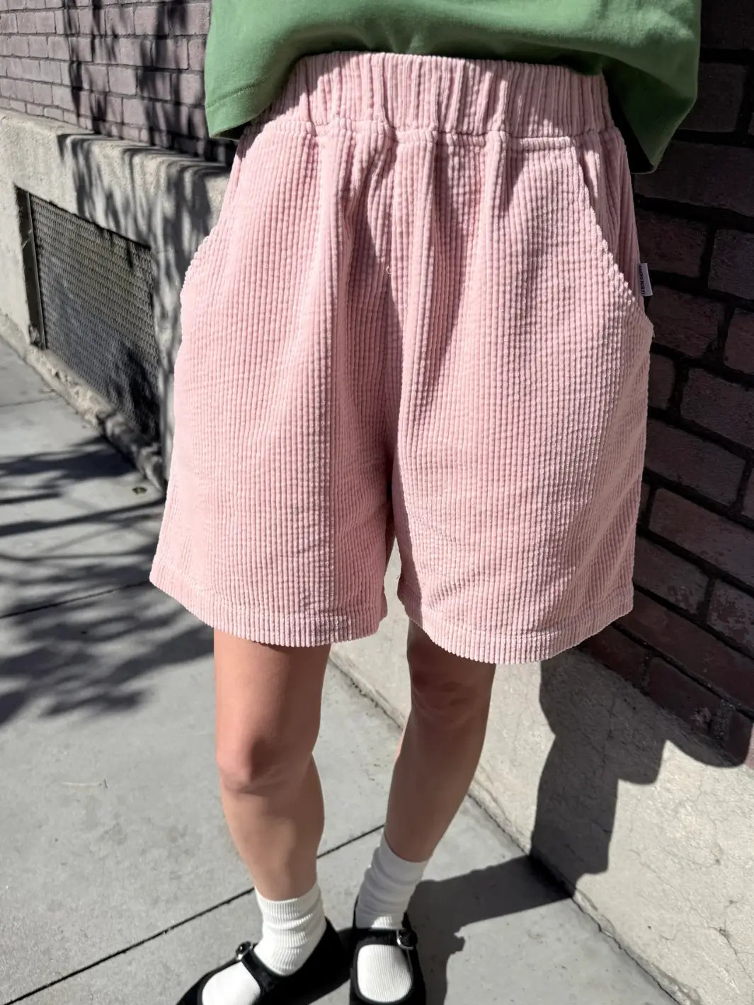 Person wearing pink 'Soft Corduroy City Shorts' in 'Baby Pink' and a green top from Le Bon Shoppe against a brick wall | Twentyseven Toronto