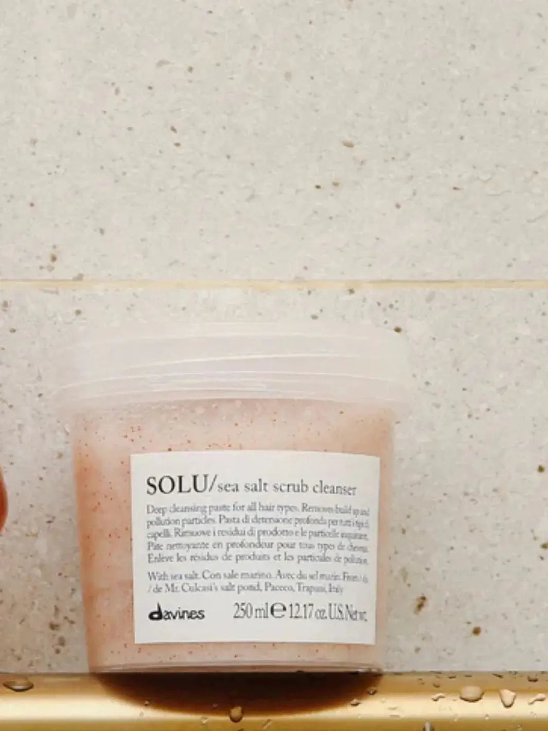 A clear tub of Davines SOLU Sea Salt Scrub Cleanser sits on a metallic gold ledge against a beige tiled wall | Twentyseven Toronto
