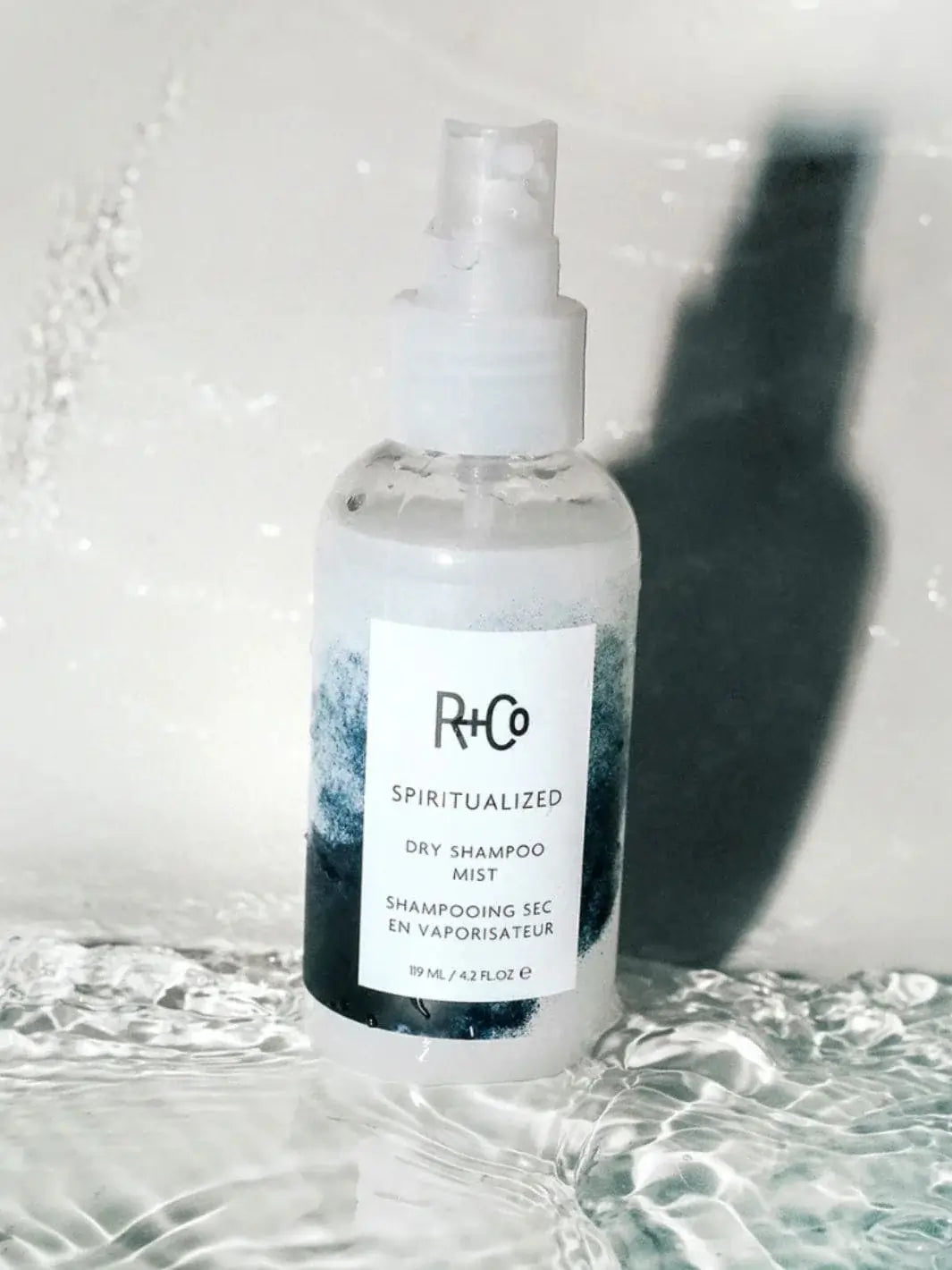 A bottle of R+Co SPIRITUALIZED Wet Dry Shampoo stands upright in a clear pool of rippling water | Twentyseven Toronto