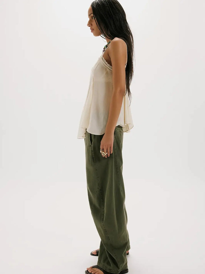 A full-length side profile view of a model wearing dark green Free People Spotlight Washed Barrel Chino Trousers in Dusty Olive paired with a flowy cream camisole and black sandals | Twentyseven Toronto
