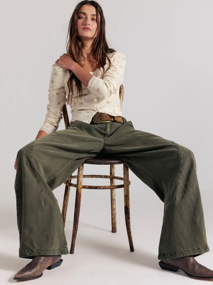 A model sits low on a rustic wooden chair, showcasing the exaggerated barrel leg shape and slouchy silhouette of the dark green Free People Spotlight Washed Barrel Chino Trousers in Dusty Olive paired with brown boots | Twentyseven Toronto