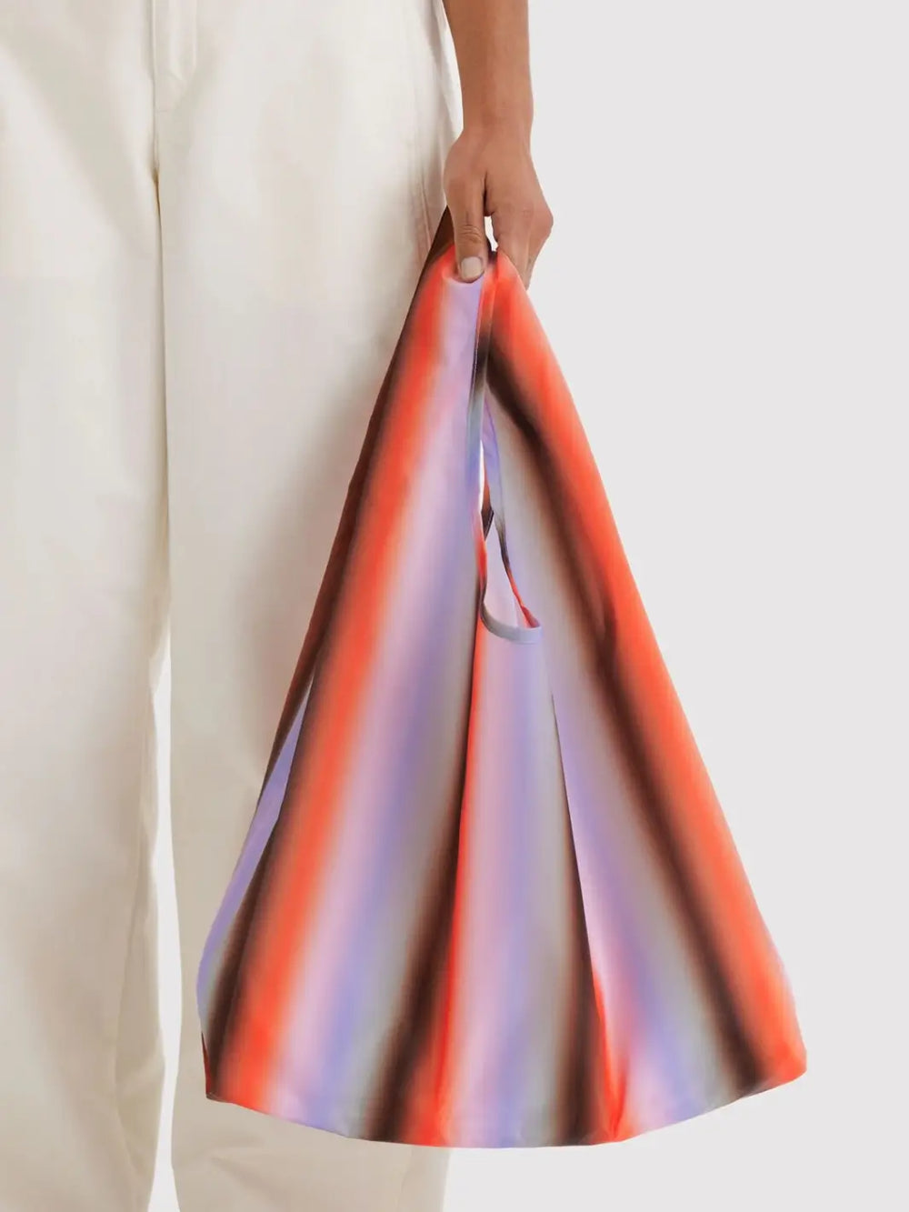 A person in white trousers holds a BAGGU Standard Baggu Bag in Gradient Stripe Poppy & Lilac | Twentyseven Toronto