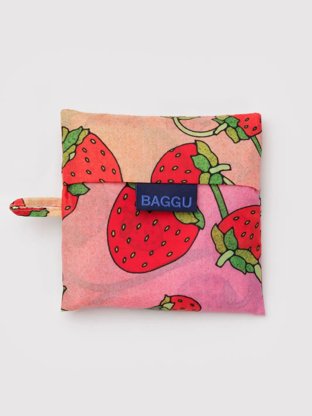 The BAGGU Standard Baggu Bag in Strawberry Blossom is shown folded into its matching square storage pouch, featuring a large-scale strawberry print and a navy blue "BAGGU" tag | Twentyseven Toronto