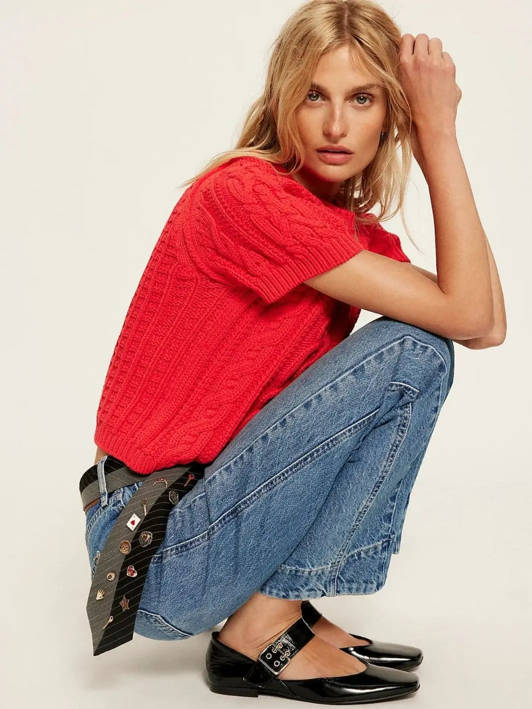 A model crouches down to show the relaxed silhouette of the Free People Stick Around Slim Curve Jeans - Stay For The Morning styled with a bright red cable-knit top and black flats | Twentyseven Toronto