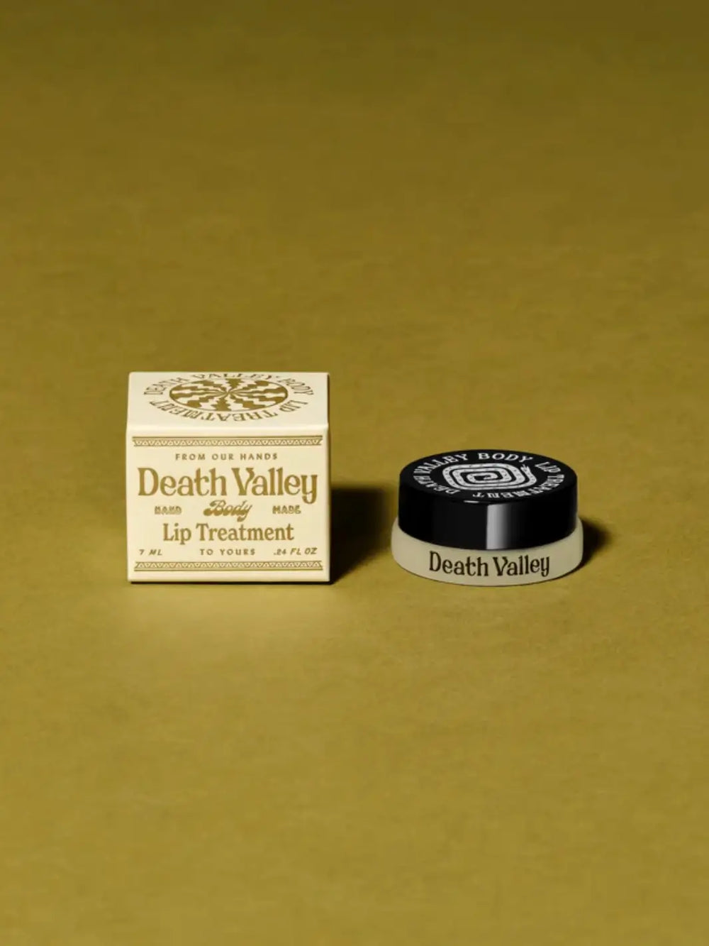 A small cosmetic pot with a black lid next to a cream-colored outer box on a green background, showcasing the packaging for Death Valley Nails Strawberry + Lime + Coconut Lip Treatment | Twentyseven Toronto