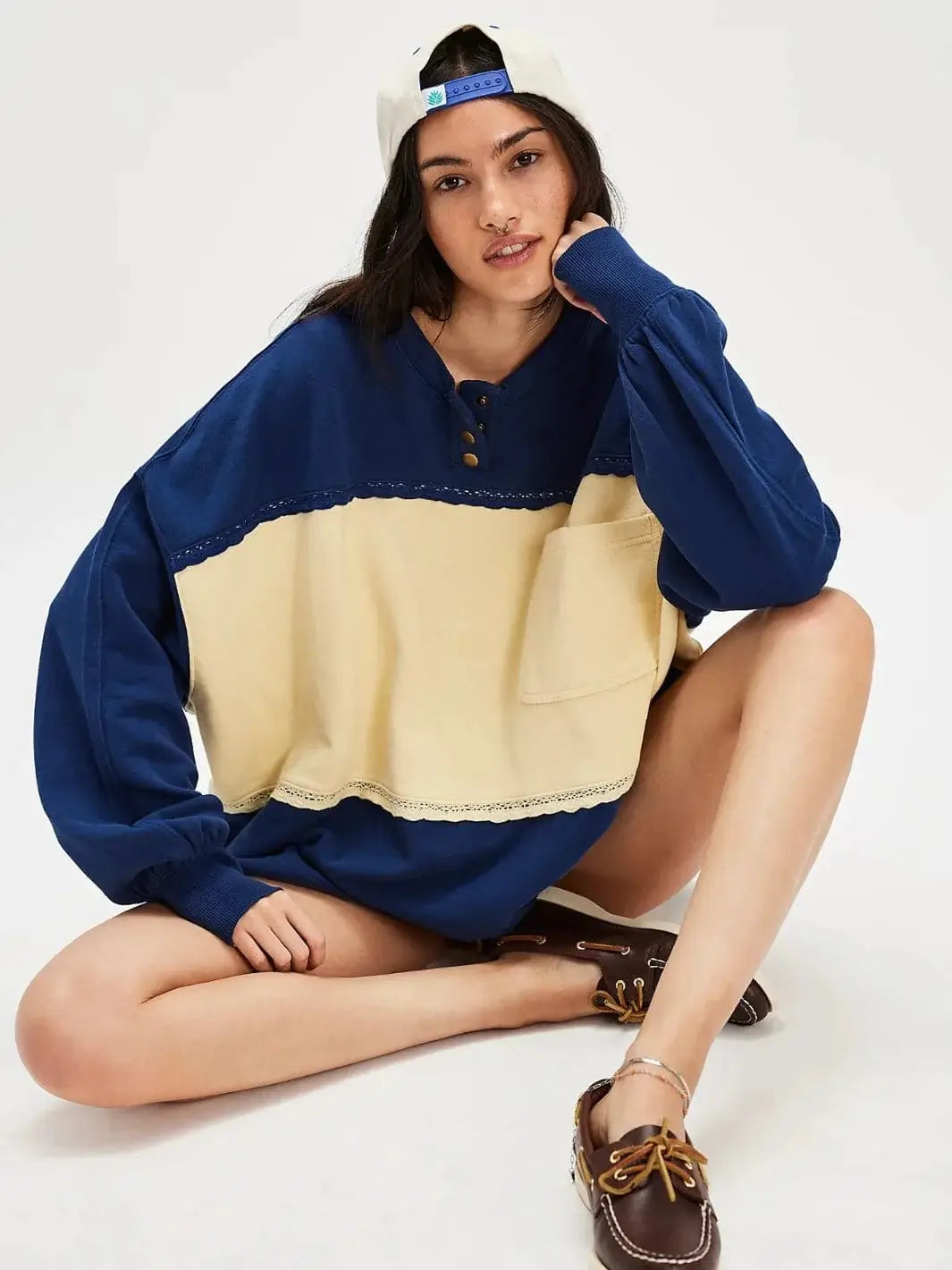 A model sits cross-legged on the floor wearing the Free People Strawberry Sweatshirt - Oceana Combo styled with a backward baseball cap and boat shoes | Twentyseven Toronto
