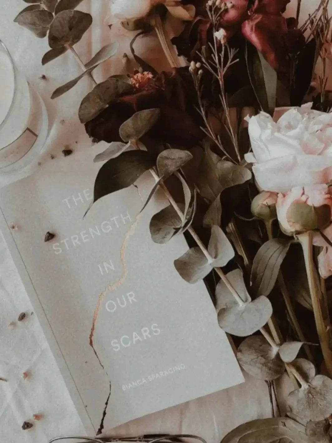 The book cover of The Strength In Our Scars by Bianca Sparacino surrounded by eucalyptus leaves and a bouquet of pink and dark red peonies | Twentyseven Toronto