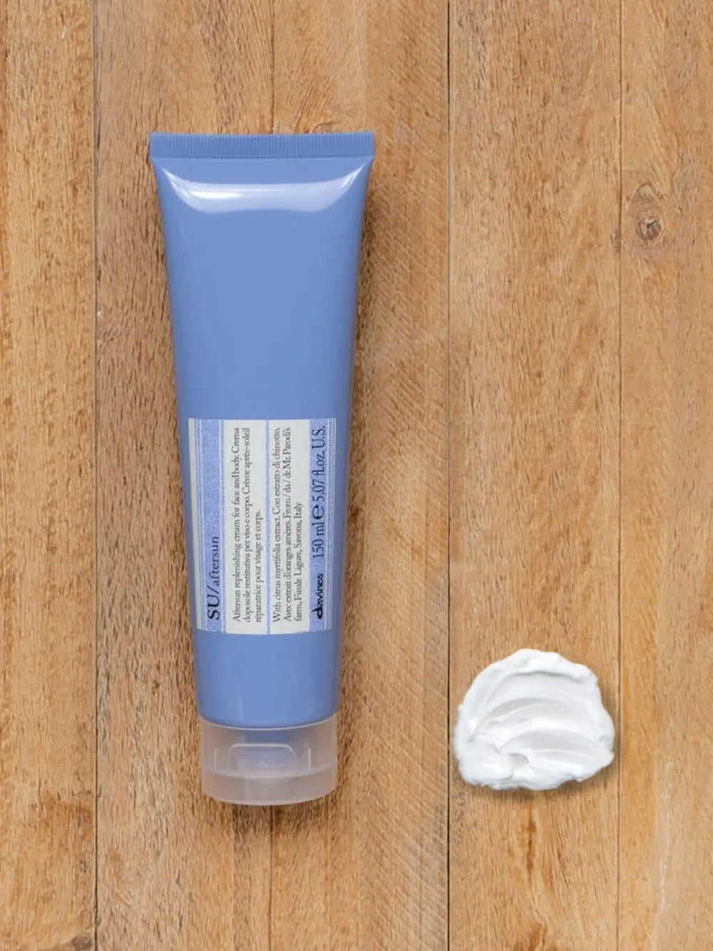 A periwinkle blue squeeze tube of Davines SU After Sun Cream for Face and Body lies vertically on a wooden surface next to a dollop of white cream | Twentyseven Toronto