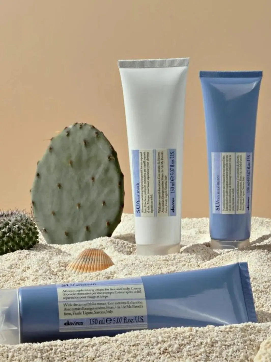 A tube of Davines SU After Sun Cream for Face and Body lies horizontally in the sand alongside other sun care products and a small cactus | Twentyseven Toronto